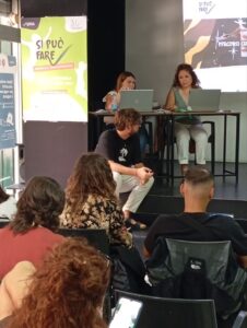 Organizzazioni no profit partecipano a un meeting del progetto 'Si Può Fare' presso uno spazio culturale. Al tavolo, due relatrici con laptop discutono dei percorsi formativi per nuove generazioni, con il pubblico attento. Sullo sfondo, cartelloni promozionali dell'evento. Iniziativa dedicata allo sviluppo delle comunità locali.